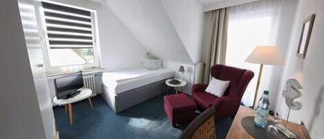 Ein kleines Hotelzimmer mit einem Bett, einem roten Sessel, einem Tisch und einem Schreibtisch.