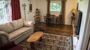 Una sala de estar con sofá, banco de madera, alfombra estampada, mesa de comedor con sillas y mueble para televisor.