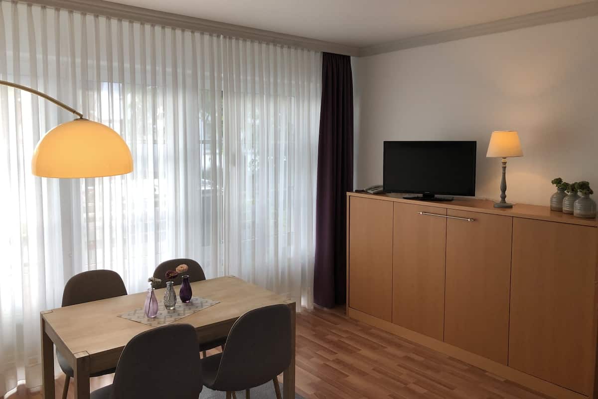 A modern hotel room with a wooden dining table, black chairs, a TV on a wooden cabinet, and a lamp.