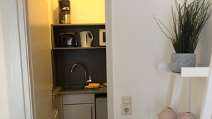 Une cuisine compacte équipée d’un évier, d’une machine à café et d’un four à micro-ondes. On y trouve également un petit placard et une plante posée sur une étagère.