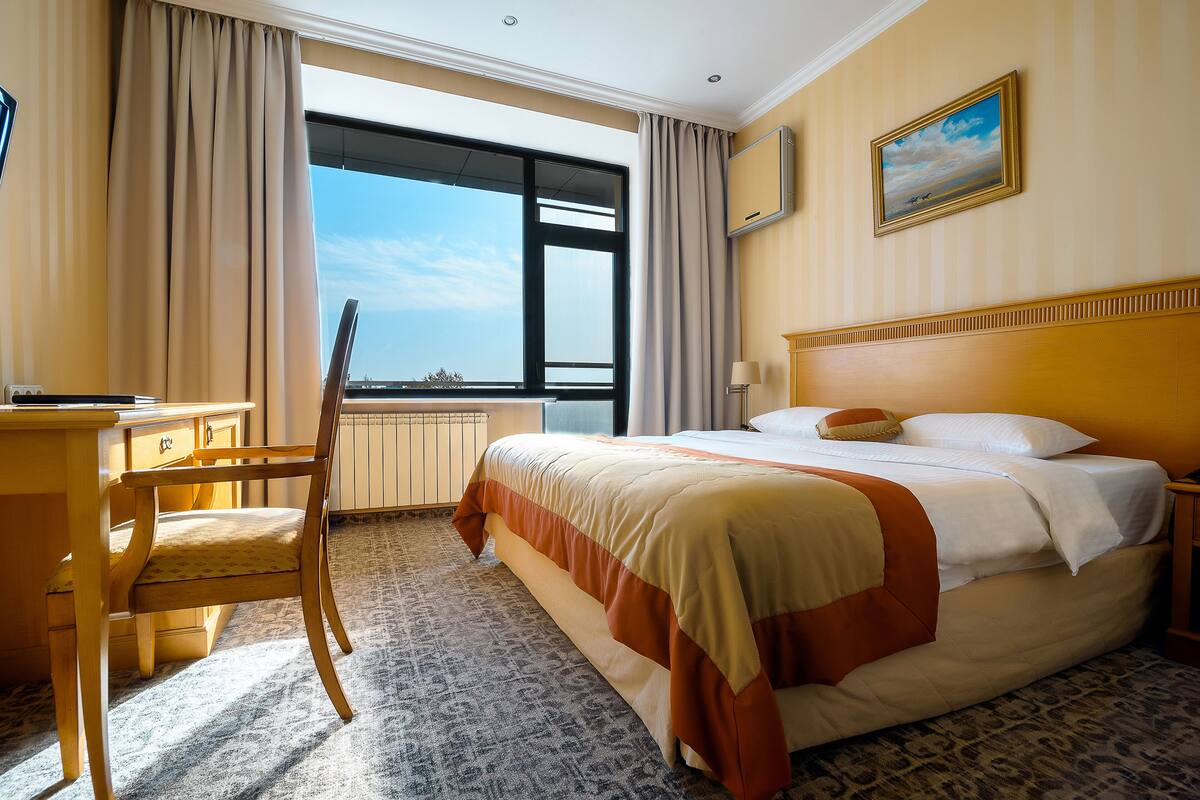 Quarto de hotel com uma cama grande, uma escrivaninha com cadeira, janela com cortinas e um quadro na parede.