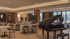 Un amplio salón de banquetes con varias mesas redondas, un piano de cola y grandes ventanales con vistas al exterior.