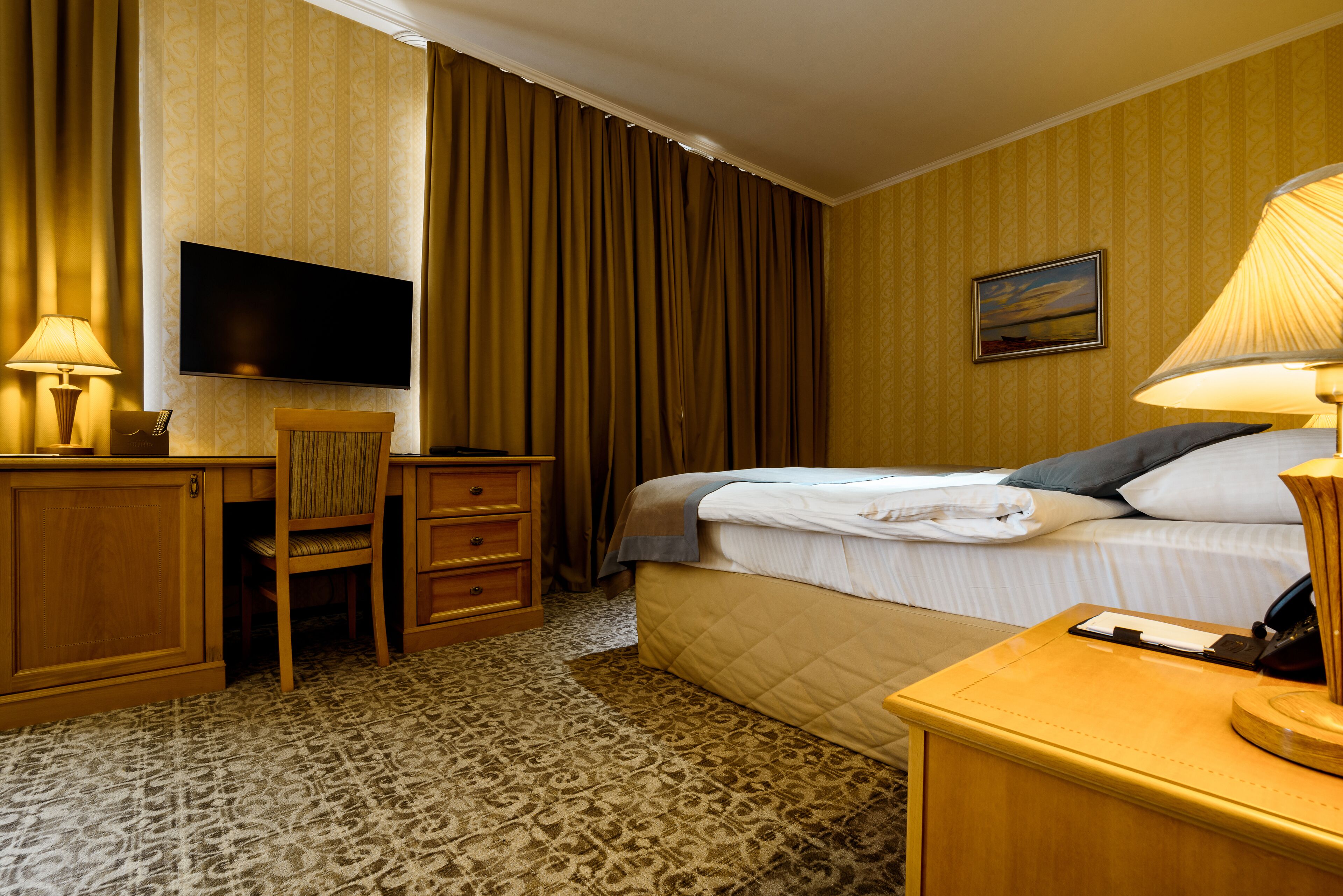 A hotel room with a bed, a desk with a chair, a television, and a painting on the wall.