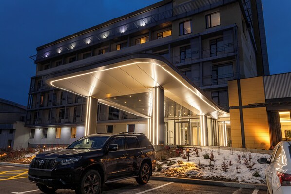Entrada de hotel moderna con un acceso cubierto, una camioneta SUV negra estacionada al frente y un coche blanco a la derecha.