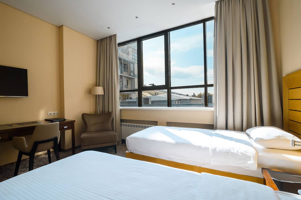 Quarto de hotel com duas camas, uma escrivaninha, uma cadeira e uma janela grande com vista para a cidade.