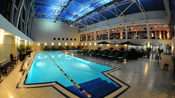 En inomhuspool med en blå vattenbana, omgiven av solstolar och krukväxter.