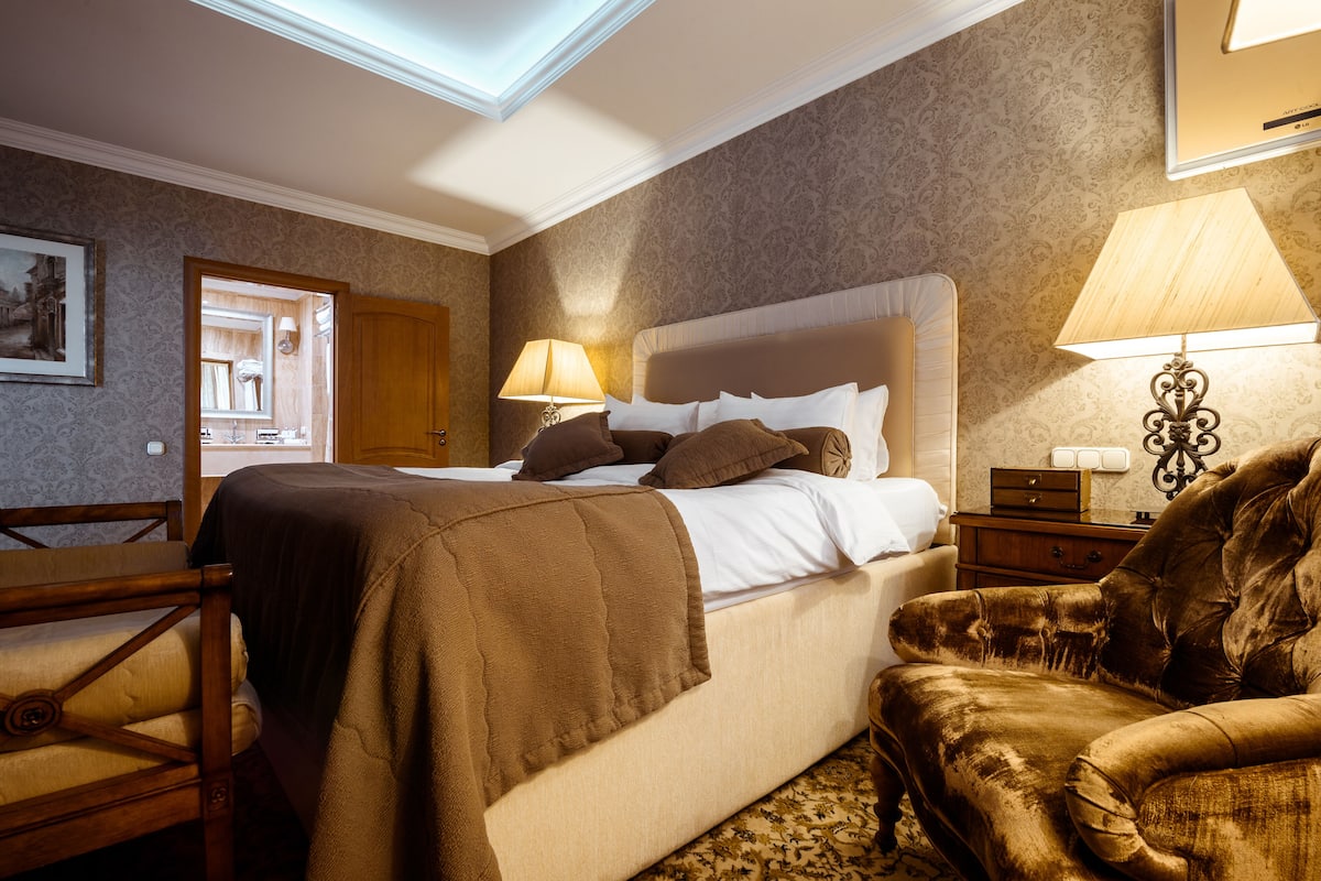 Quarto de hotel com uma cama grande, uma cadeira de madeira, uma mesa de cabeceira e um abajur.