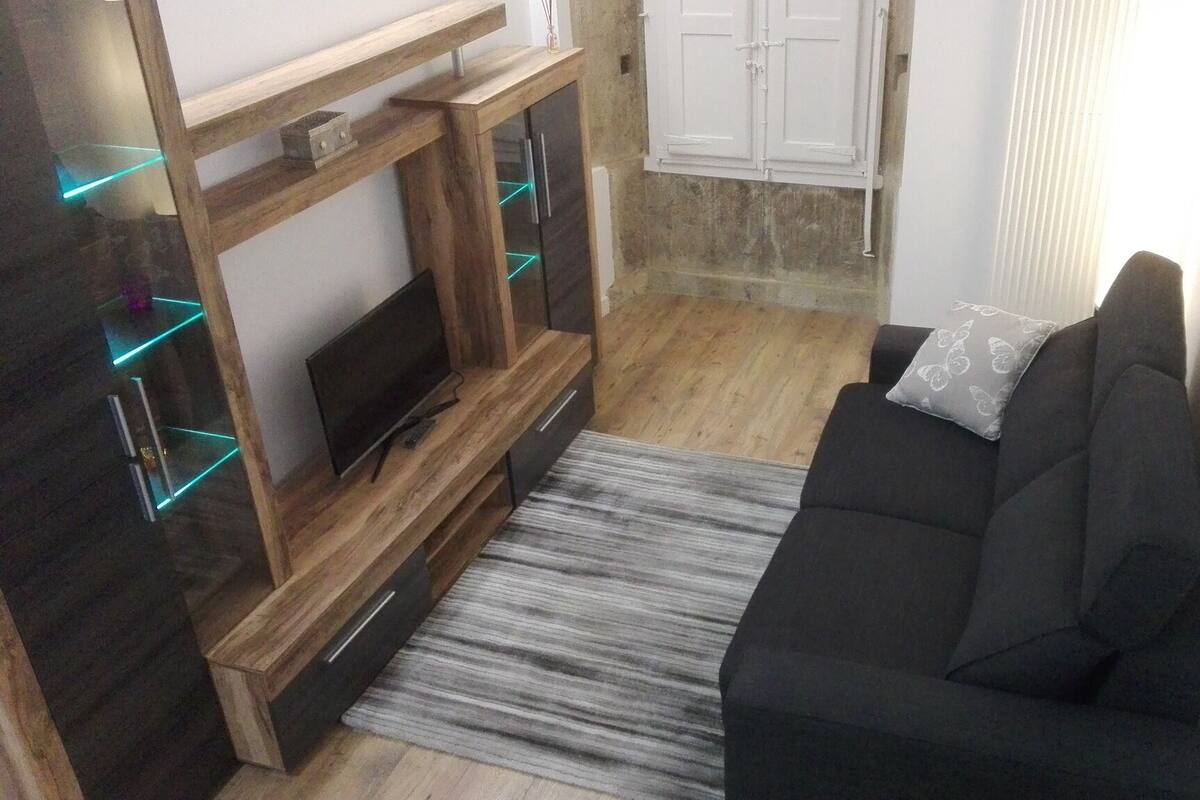 A living room with a dark sofa, a wooden TV stand with a flat-screen TV, and a patterned rug.