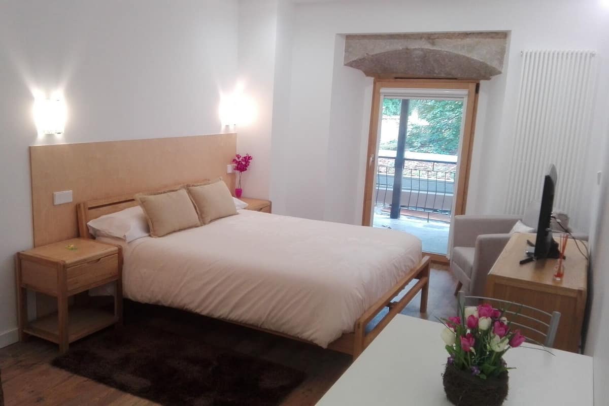 A bedroom with a bed, bedside tables, a desk with a computer, a chair, a window with a view, and a small plant.