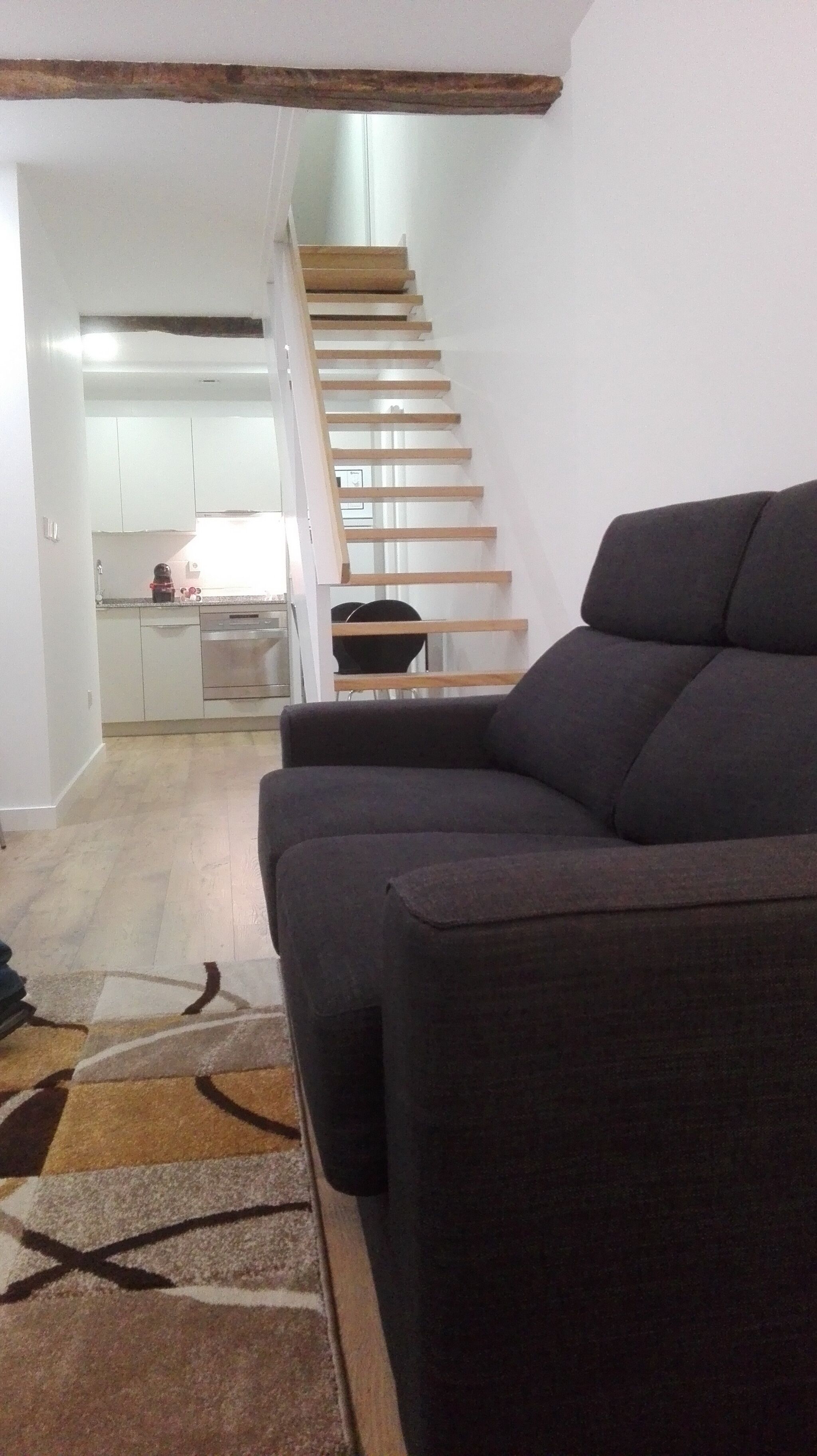 Ein Wohnzimmer mit einem dunklen Sofa, einer Treppe, die zu einer Küche führt, und einem gemusterten Teppich.