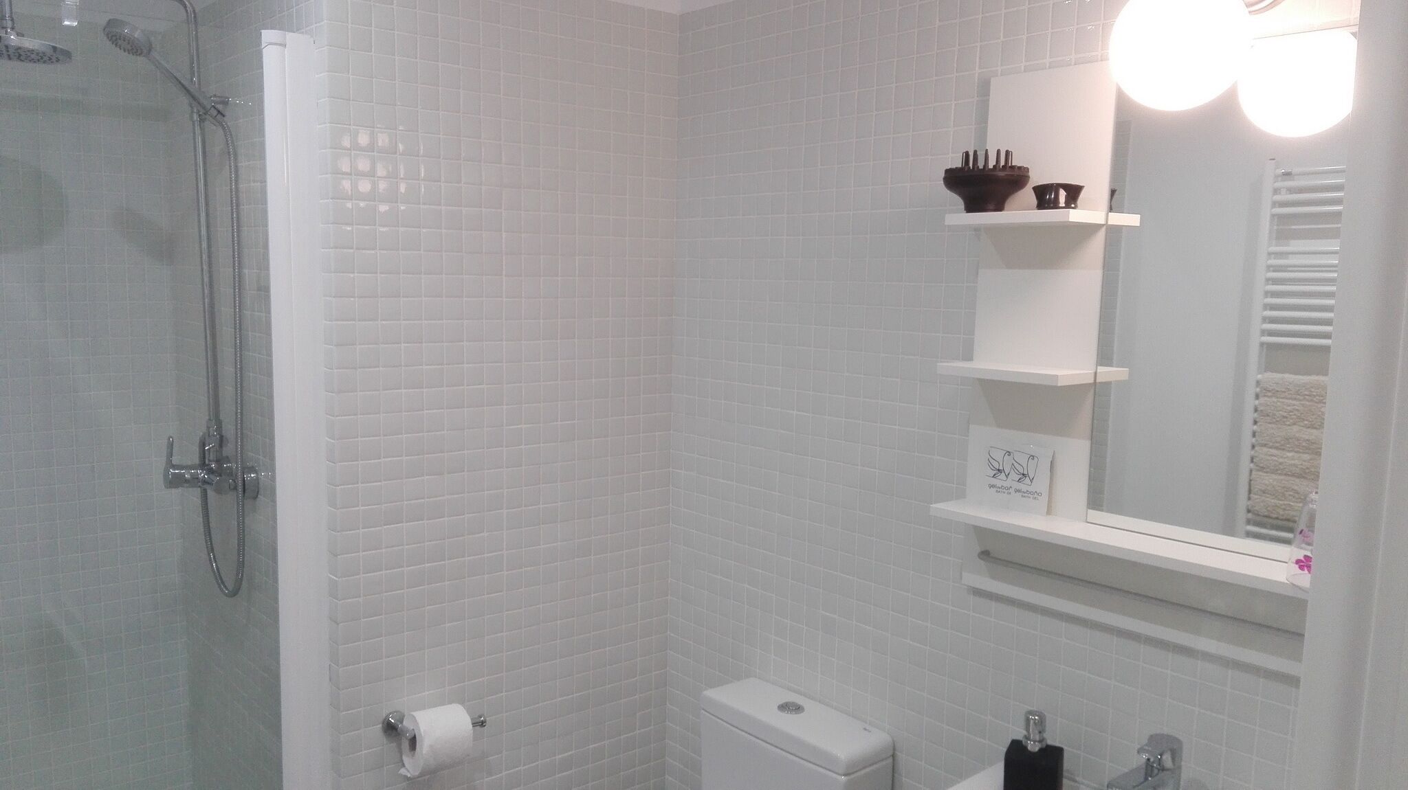 Une salle de bain avec des murs recouverts de carreaux blancs, une douche, des toilettes, un lavabo et un miroir avec un éclairage au-dessus.