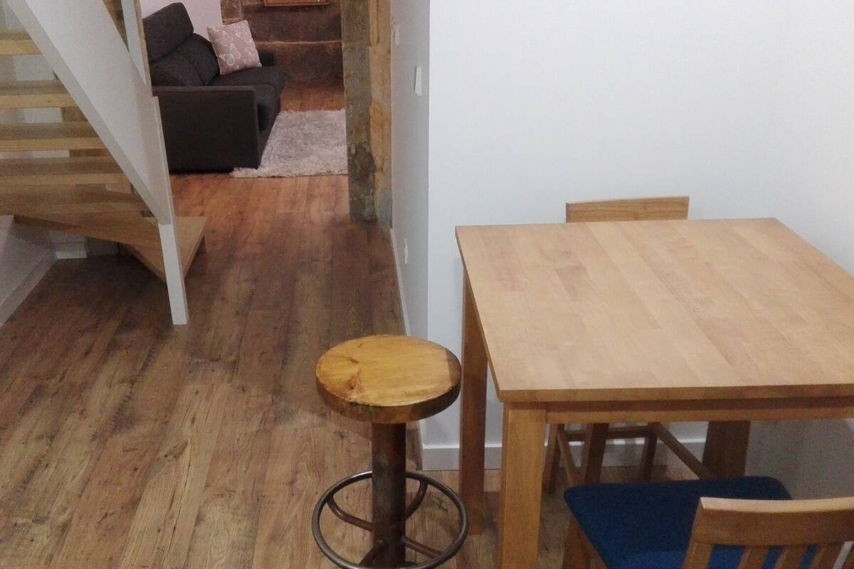 A wooden dining table with chairs, a small stool, and a staircase leading to a living area with a sofa and cushions.