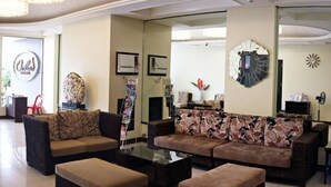Amplia sala de estar con un sofá grande, dos otomanas y una mesa de centro. Se observa un espejo grande en la pared, un reloj y un adorno mural.