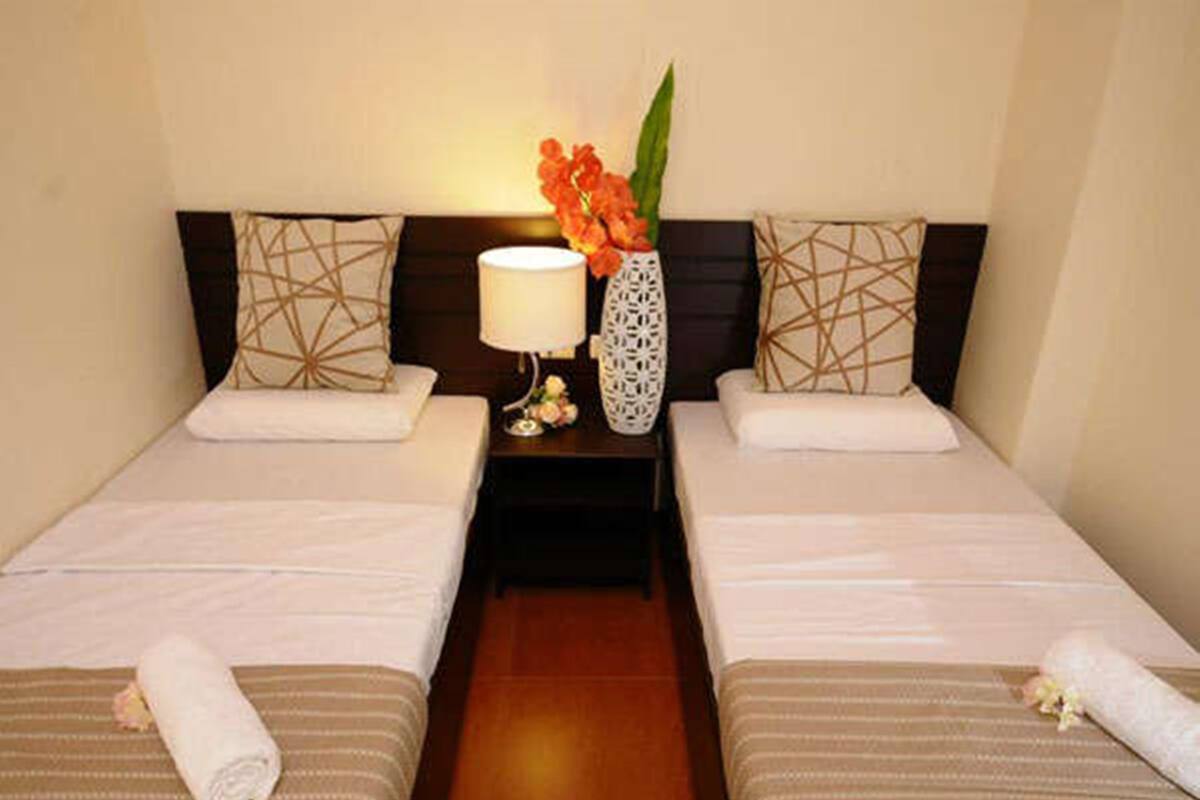 A hotel room with two beds, a nightstand with a lamp, a vase with flowers, and patterned pillows.