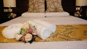 Una cama bien hecha con toallas blancas enrolladas y un arreglo floral sobre un cubrecama tejido.