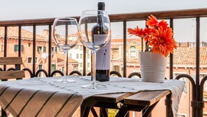 A table with two wine glasses and a bottle of wine, a potted orange flower, and a view of buildings in the background.