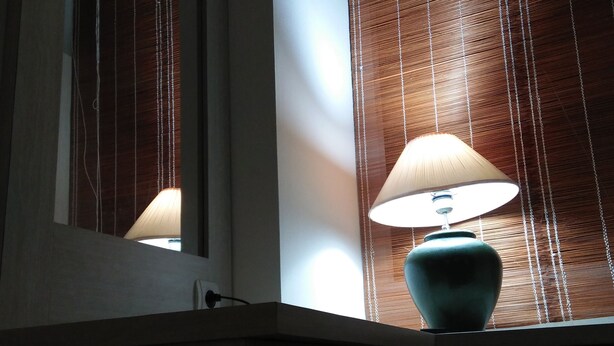 A bathroom with a mirror, a window with blinds, and a lamp on a counter. - Shamrock Vivo Apartemen (Depok)