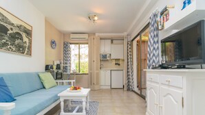 A modern living room with a blue sofa, a small white table with fruit, a TV, and a kitchen area with cabinets and a microwave. - ENCINAS 1 - Apartment with terrace in Port De Pollença. Free WiFi (Pollença)