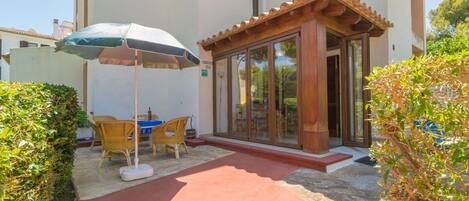 A covered outdoor seating area with a table and chairs, a wooden pergola, and a glass door entrance to a building.
