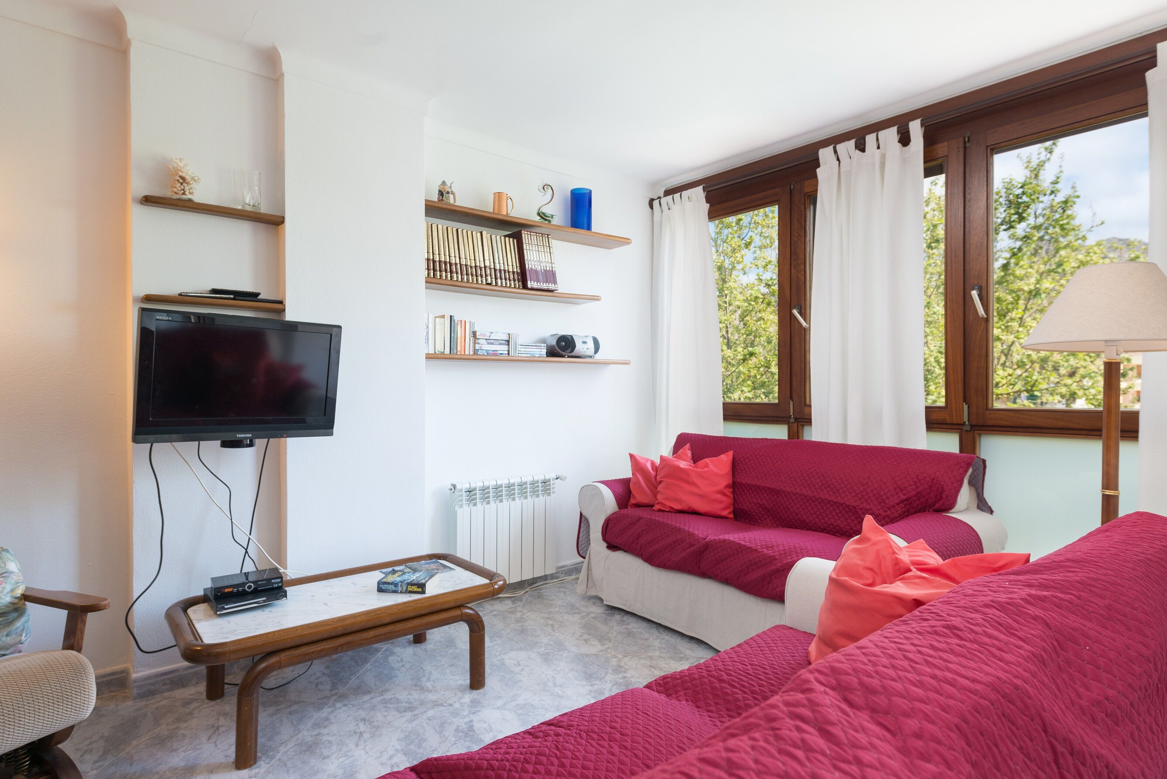 A living room with a TV, shelves with books and decorative items, a red sofa, and a window with curtains.