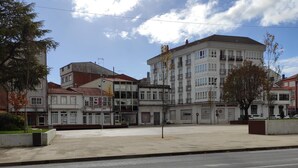A multi-story building with a commercial ground floor, featuring a variety of storefronts and a parking lot in front. - Albergue Arraigos - Hostel (Melide)