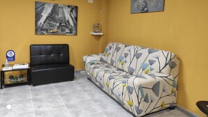 A room with a yellow wall, a black sofa, a patterned couch, a black leather chair, a small table with a bottle and papers, and a framed picture on the wall. - Albergue Arraigos - Hostel (Melide)