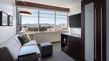 A modern living room with a sofa, ottoman, and a TV on a stand. There is a window with a city view, a cabinet with shelves, and framed pictures on the wall.