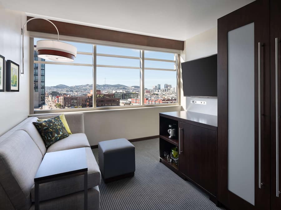 A modern living room with a sofa, ottoman, and a TV on a stand. There is a window with a city view, a cabinet with shelves, and framed pictures on the wall.