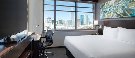 A hotel room with a large bed, a desk, a chair, and a window with a city view.