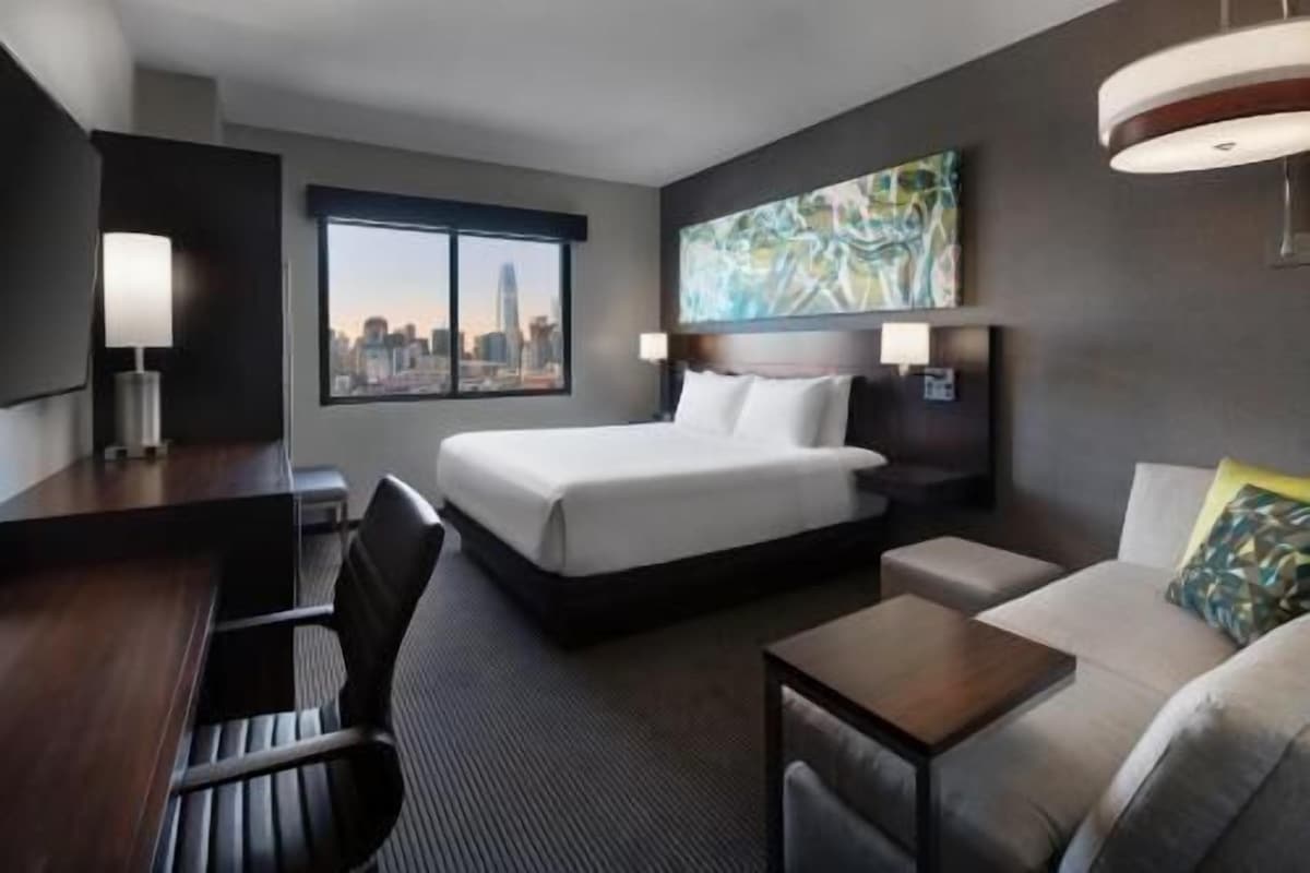 A hotel room with a bed, a desk, a chair, a sofa, a window with a city view, and a painting on the wall.
