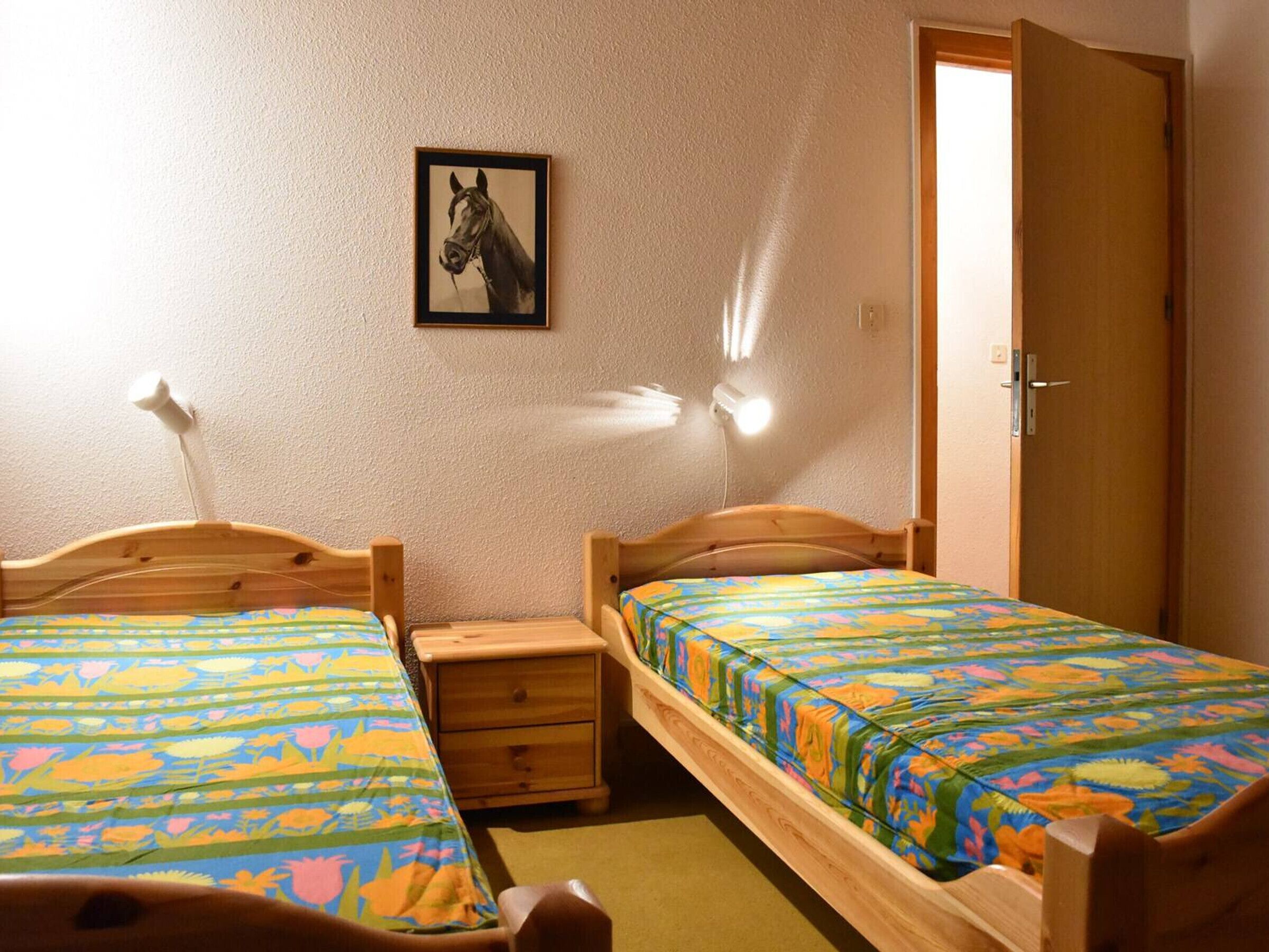 Una habitación con dos camas de madera, una mesita de noche, un cajón pequeño, un cuadro enmarcado en la pared y una puerta.