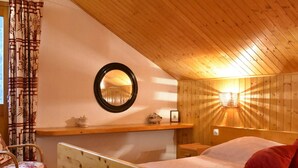 Une chambre confortable avec un plafond en bois, un miroir rond, un lit avec une literie blanche et un oreiller rouge, une table de chevet en bois et une chaise en osier.