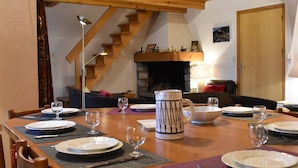 Un coin repas avec une table en bois dressée pour quatre personnes, une cheminée et un escalier en arrière-plan.