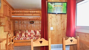 Una habitación acogedora de madera con una cama, un televisor montado en la pared y un ventanal con cortinas rojas.