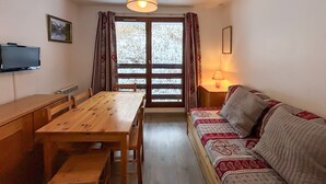 A cozy room with a wooden dining table, a sofa, a TV, and a window with curtains. - Economical studio for 3 in the heart of Les Menuires (Les Menuires)