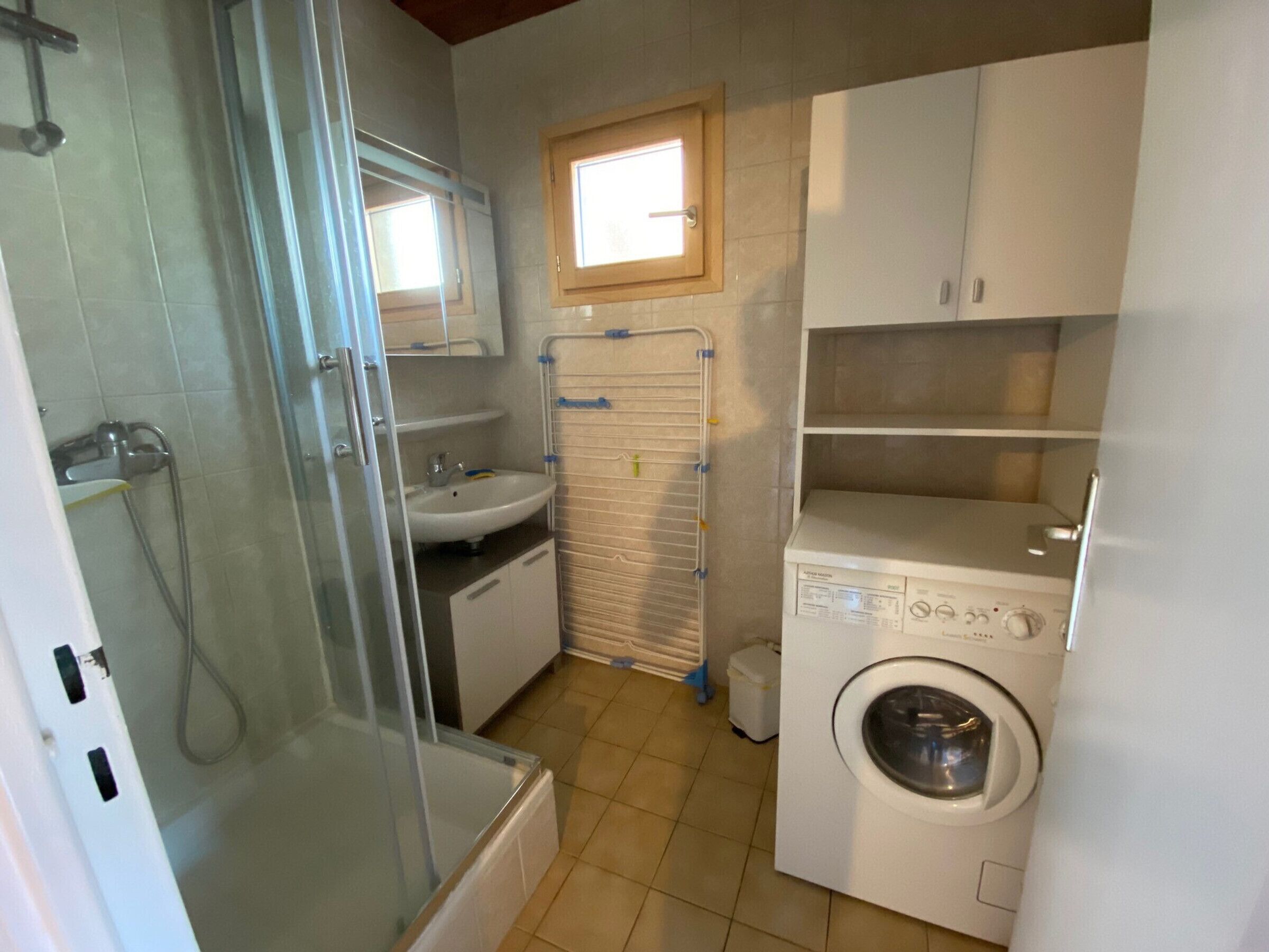 A compact bathroom with a glass shower enclosure, a sink, a mirror, a window, and a washing machine.