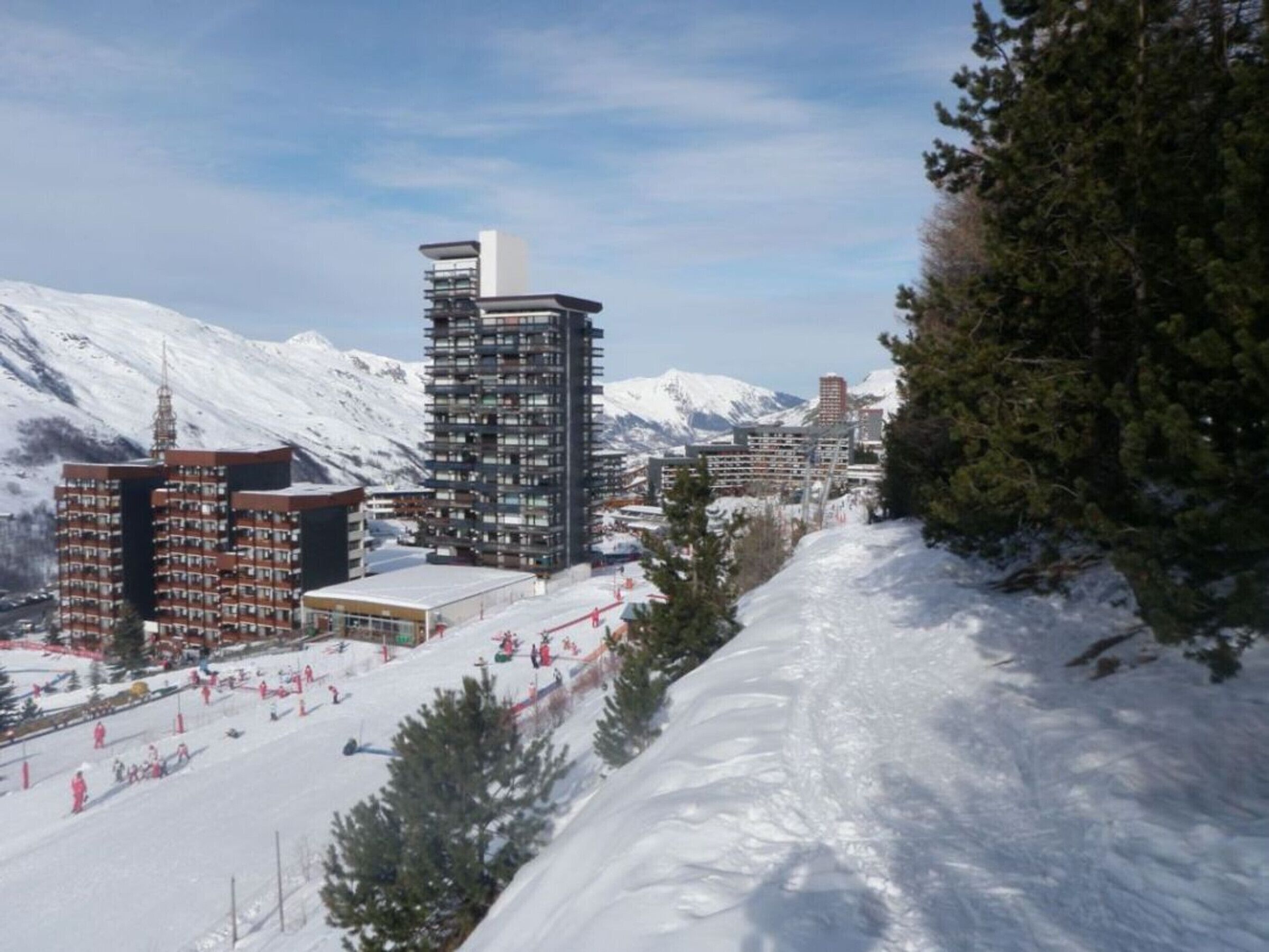 A ski resort with modern buildings, including a tall hotel with a unique rooftop design, surrounded by snow-covered mountains.