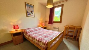 A bedroom with a wooden bed, a nightstand with a lamp, a radiator, a window with a view, and a poster on the wall.