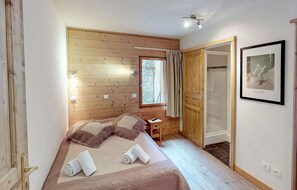 Quarto com teto de madeira, cama com toalhas dobradas, mesa de cabeceira, quadro emoldurado e janela com cortinas.