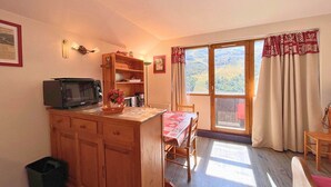 Une cuisine compacte équipée d’un four à micro-ondes, de meubles en bois et d’un coin repas avec une table et des chaises. Une fenêtre avec des rideaux offre une vue sur l’extérieur.