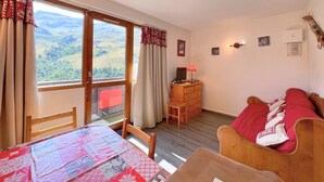 Une pièce chaleureuse avec un plancher en bois, un lit, un bureau avec une lampe, une chaise et une vue sur la campagne grâce à de grandes fenêtres.
