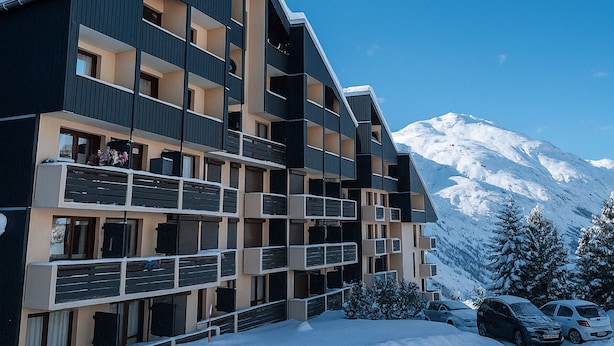 Un hôtel à plusieurs étages doté de balcons, avec une montagne enneigée en arrière-plan et des voitures garées.