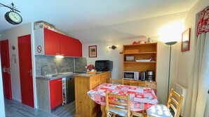 Une cuisine de taille réduite avec des meubles rouges, un évier et un four à micro-ondes. On y trouve une table à manger avec des chaises et une horloge au mur.
