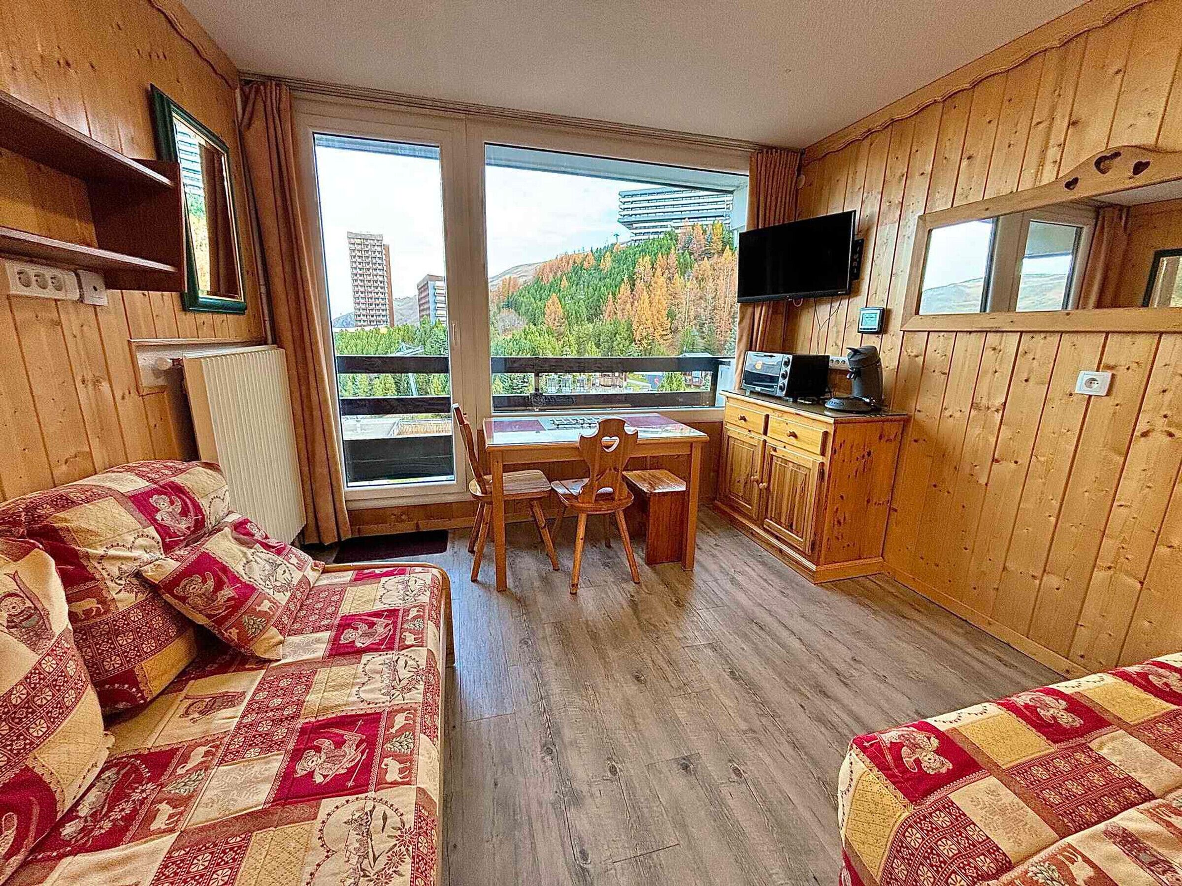 A cabin with wooden walls, a dining area, a TV, and a view of a forest and buildings.