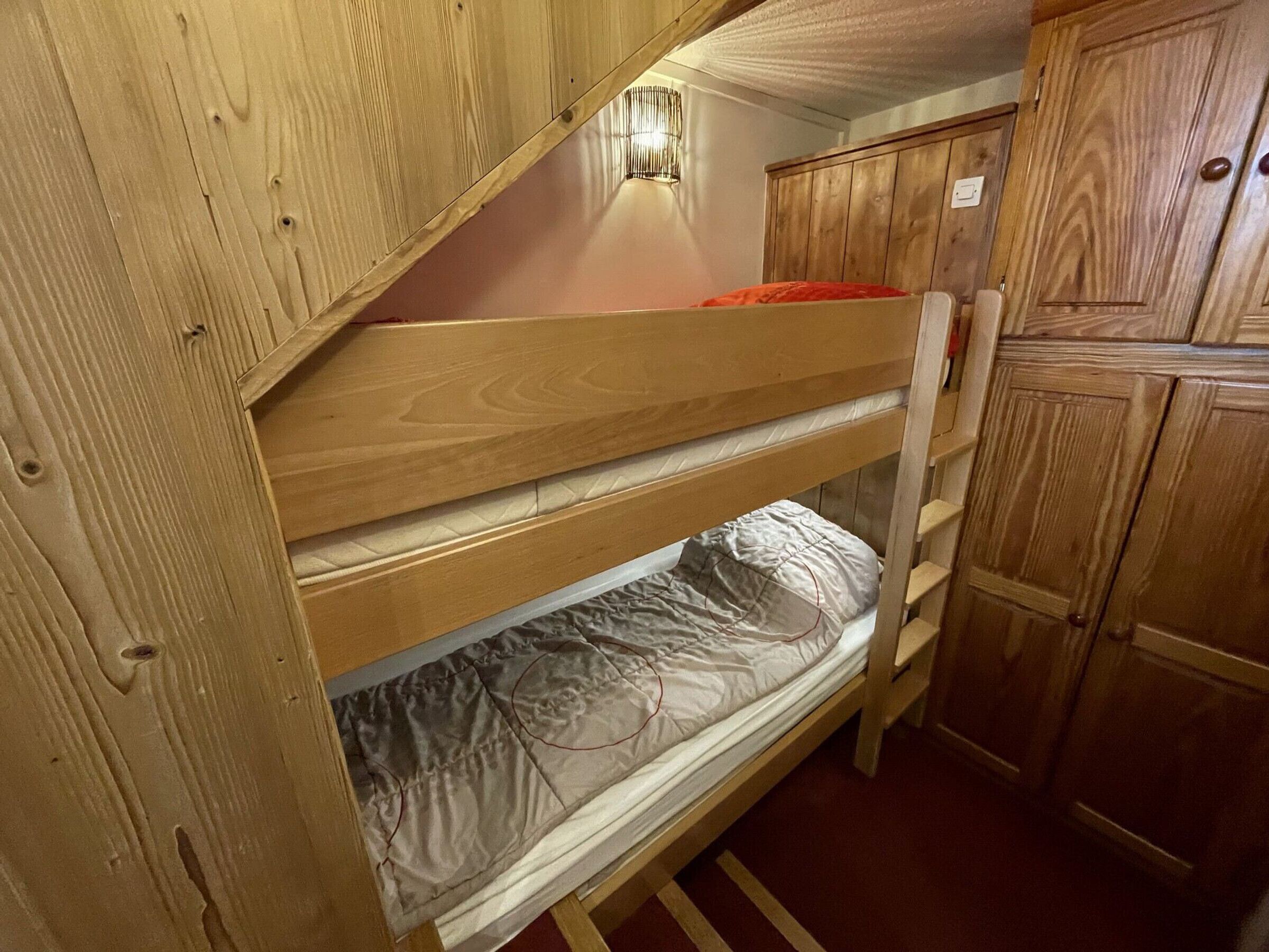 Una cama litera con cabecera y piecera de madera, un almohón rojo y un colchón blanco.