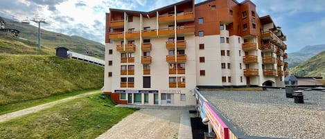 A multi-story hotel with balconies, a ski lift in the background, and a gravel path leading to the entrance.