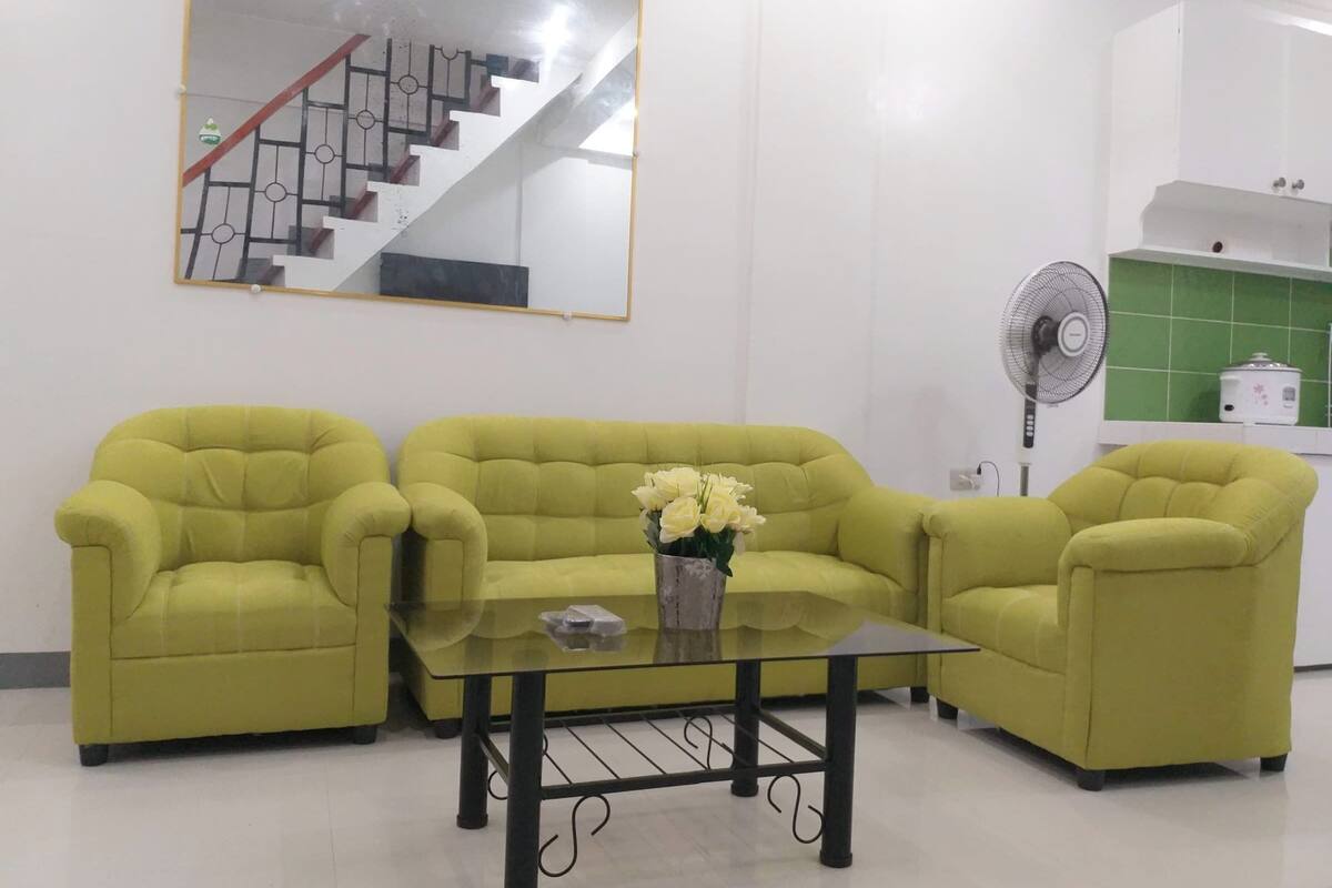 A living room with a yellow sofa set, a glass coffee table, and a white kitchen area with a fan and a rice cooker.