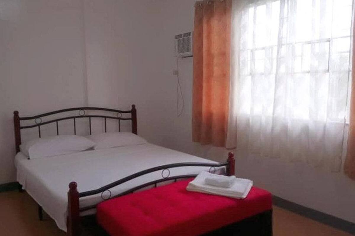 A single bed with a wooden frame, a white mattress, and a red blanket. There is a window with curtains, and an air conditioning unit mounted on the wall.