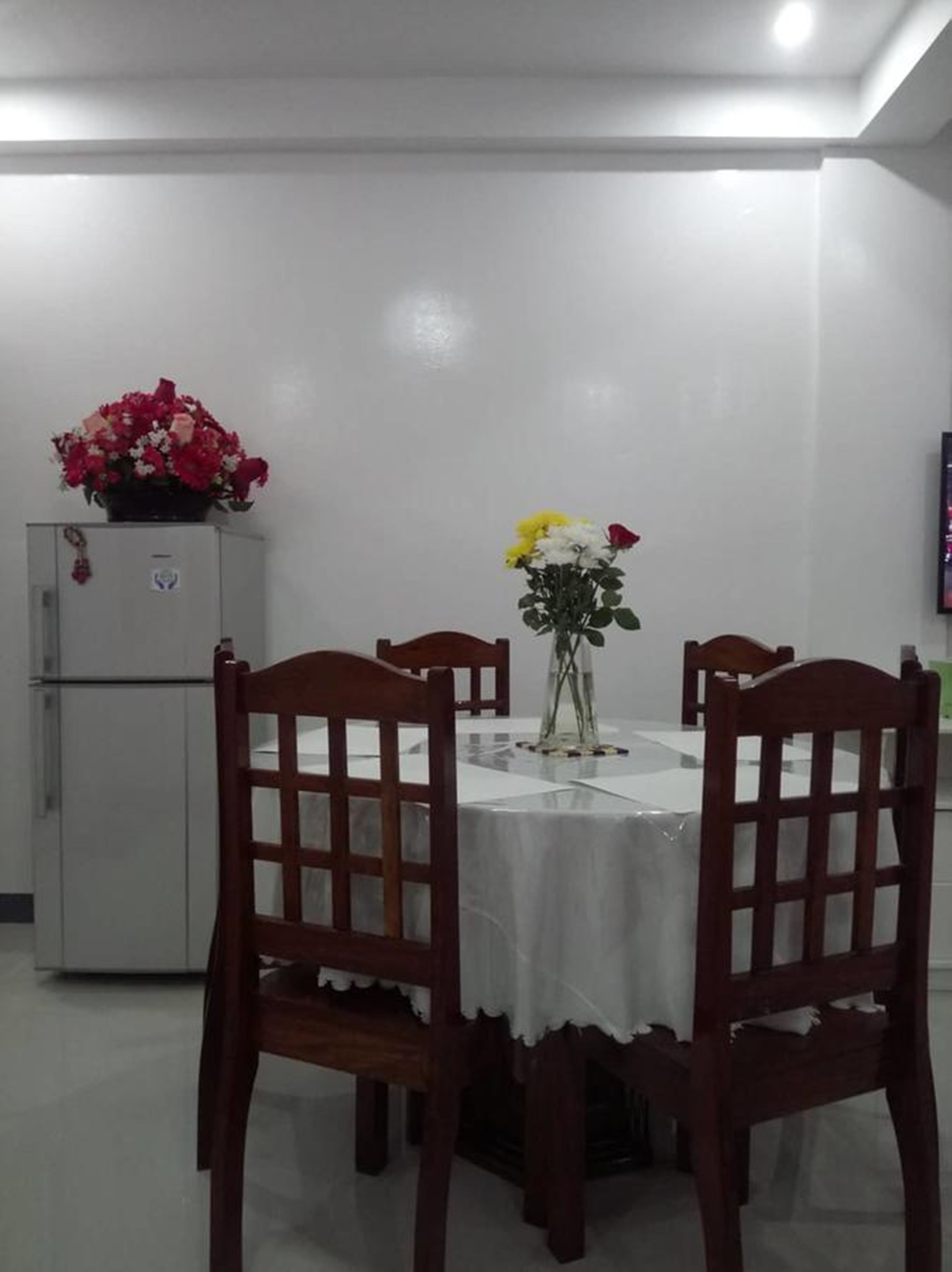 A dining area with a round table covered by a white tablecloth, surrounded by wooden chairs with a grid pattern. A refrigerator with a floral decoration is visible in the background.