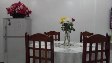 A dining area with a round table covered by a white tablecloth, surrounded by wooden chairs with a grid pattern. A refrigerator with a floral decoration is visible in the background.